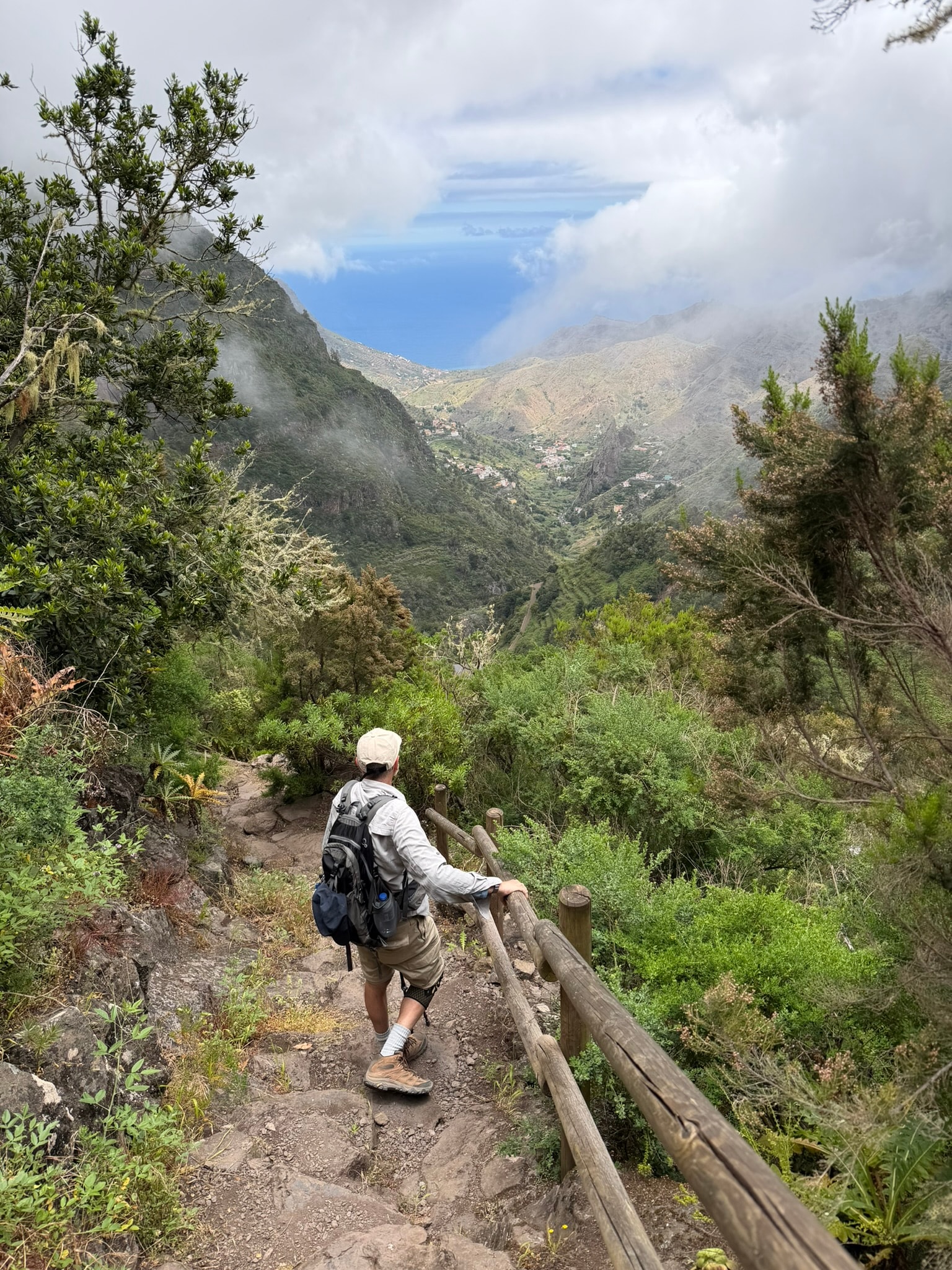 A hiker descending a mountain trail with a wide green valley and distant peaks stretching below