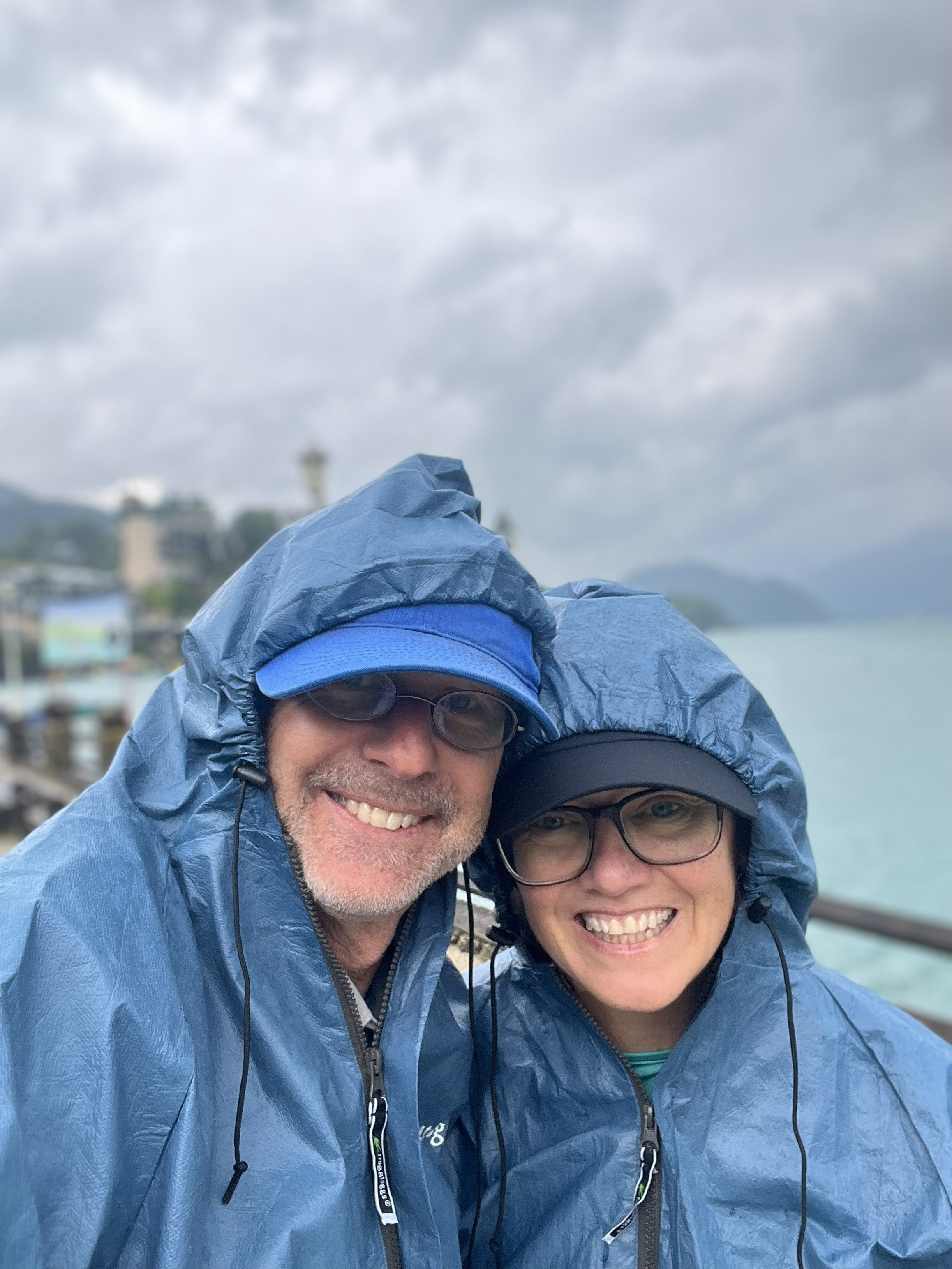 Cheryl and Steve smiling in matching blue rain jackets at a lakeside waterfront