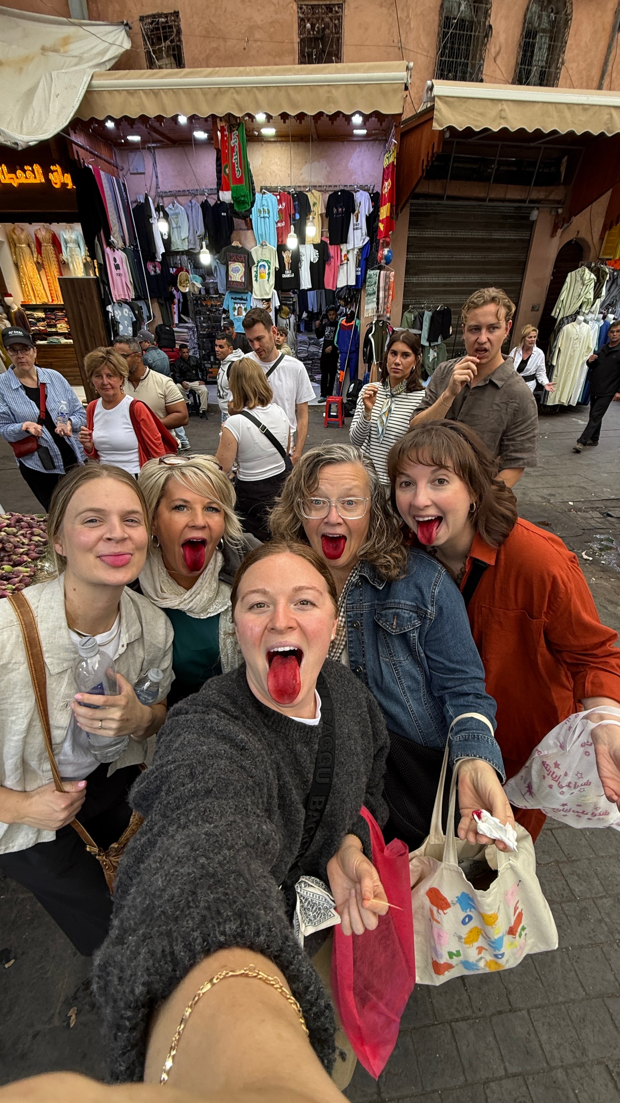 Five women in the Marrakech souk sticking out their red-stained tongues after tasting something from a market stall, laughing in front of clothing shops