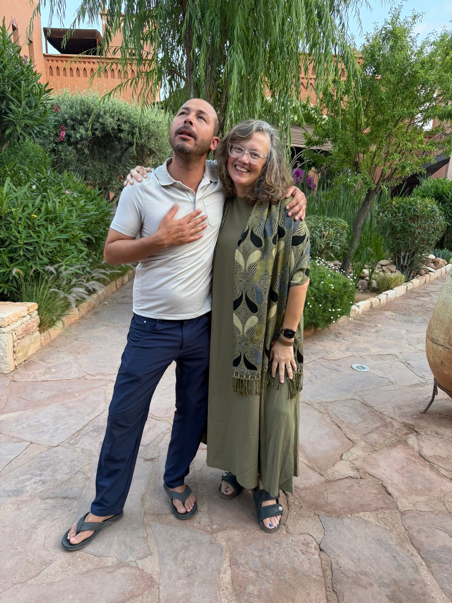 Cheryl and Fouad embracing in a sunlit Moroccan garden courtyard, both smiling warmly