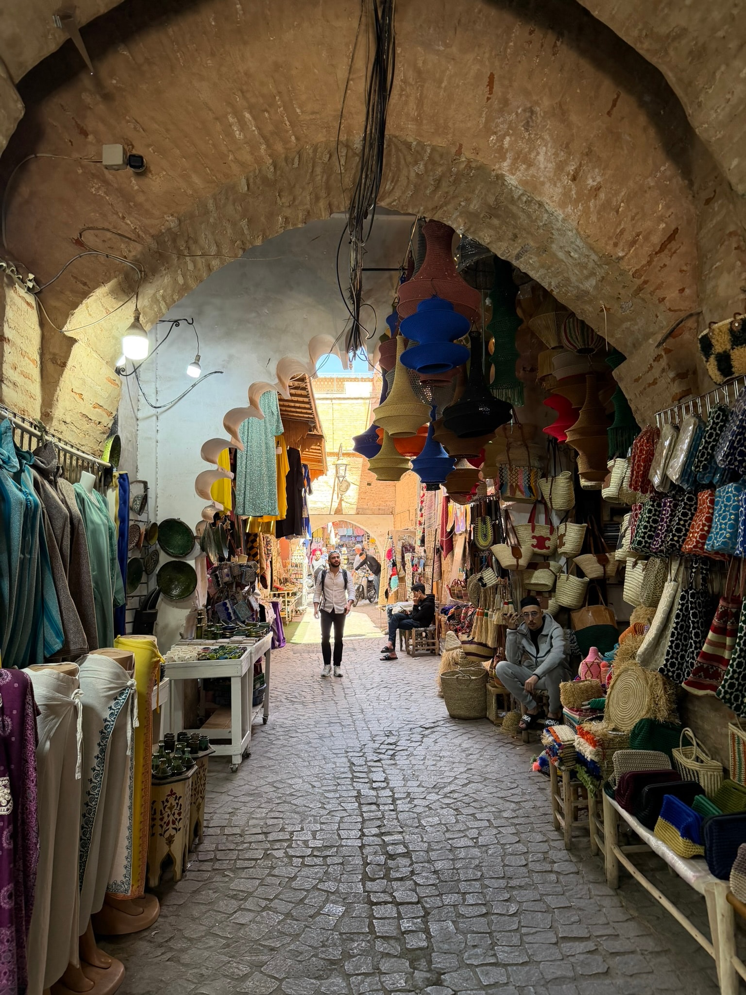 A cobblestone lane in the Marrakech medina, arched overhead with stone, lined on both sides with hanging tagines, woven baskets, and colorful textiles