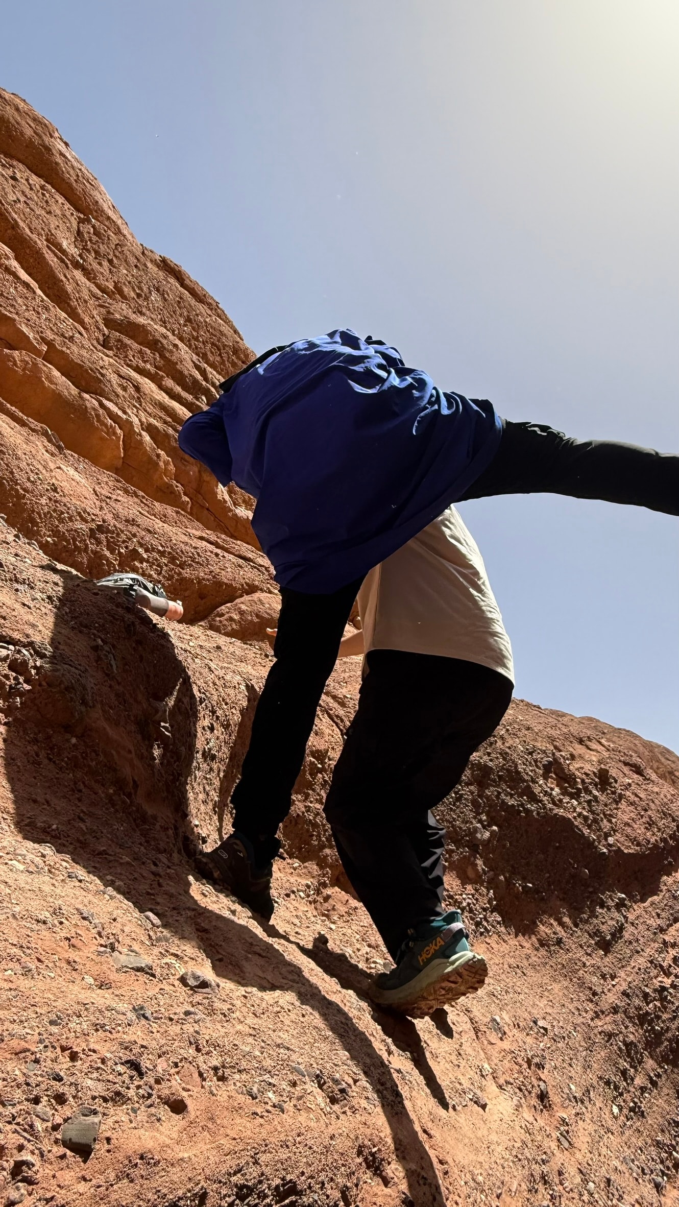 Person rock-climbing a steep sandstone face in the Atlas Mountains, arms spread wide for balance against a bright sky