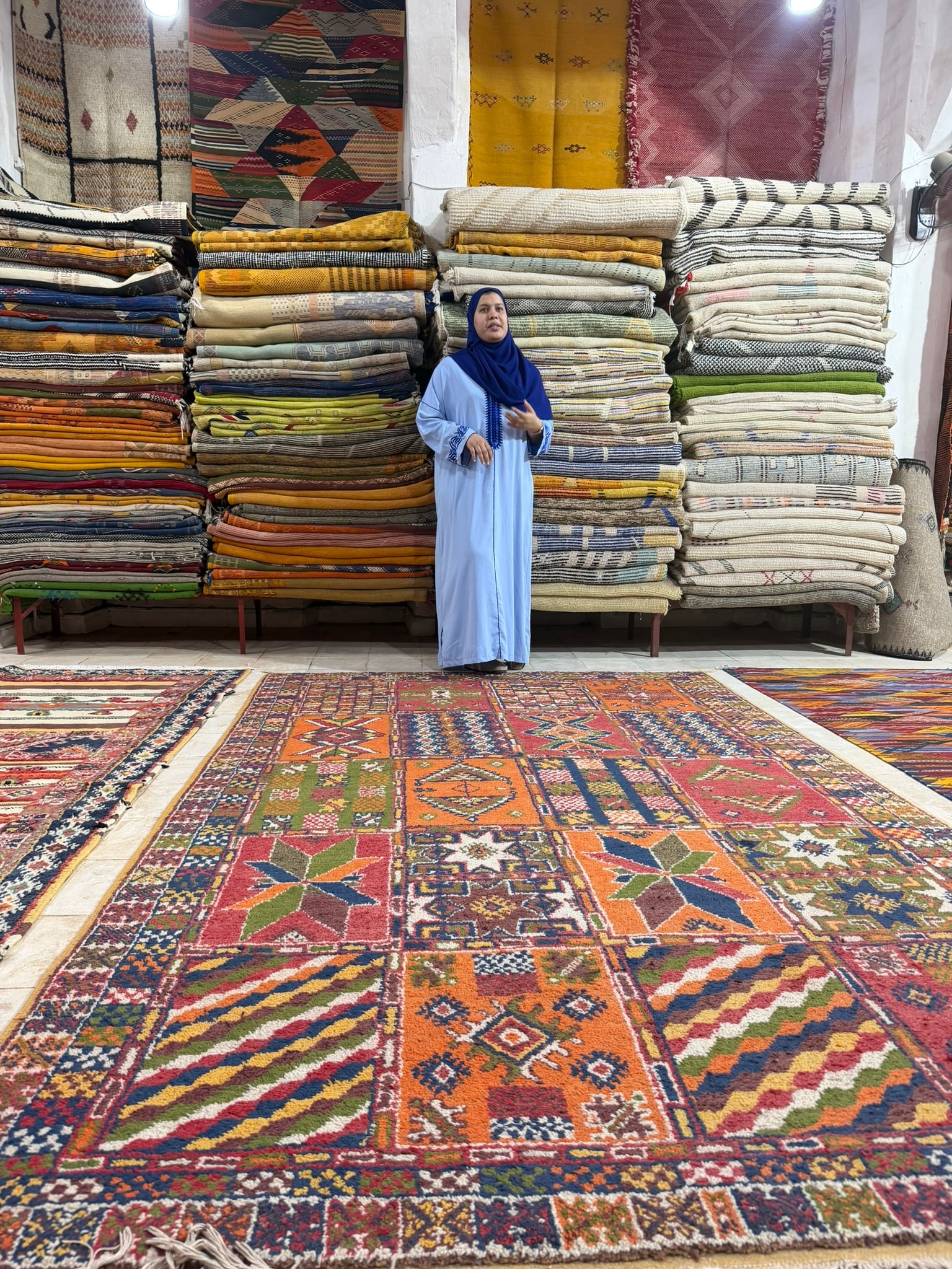A woman in a light blue djellaba and hijab stands surrounded by floor-to-ceiling stacks of colorful handwoven Moroccan rugs and kilims in a cooperative workshop