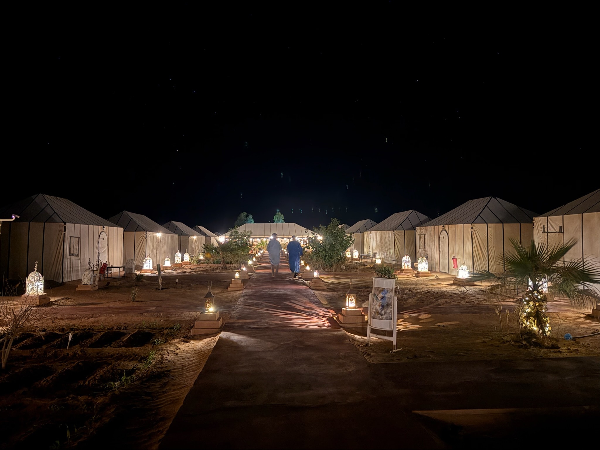 Two figures in robes walking down a lantern-lit pathway between rows of white glamping tents under a star-filled sky at the Sahara desert camp, Morocco