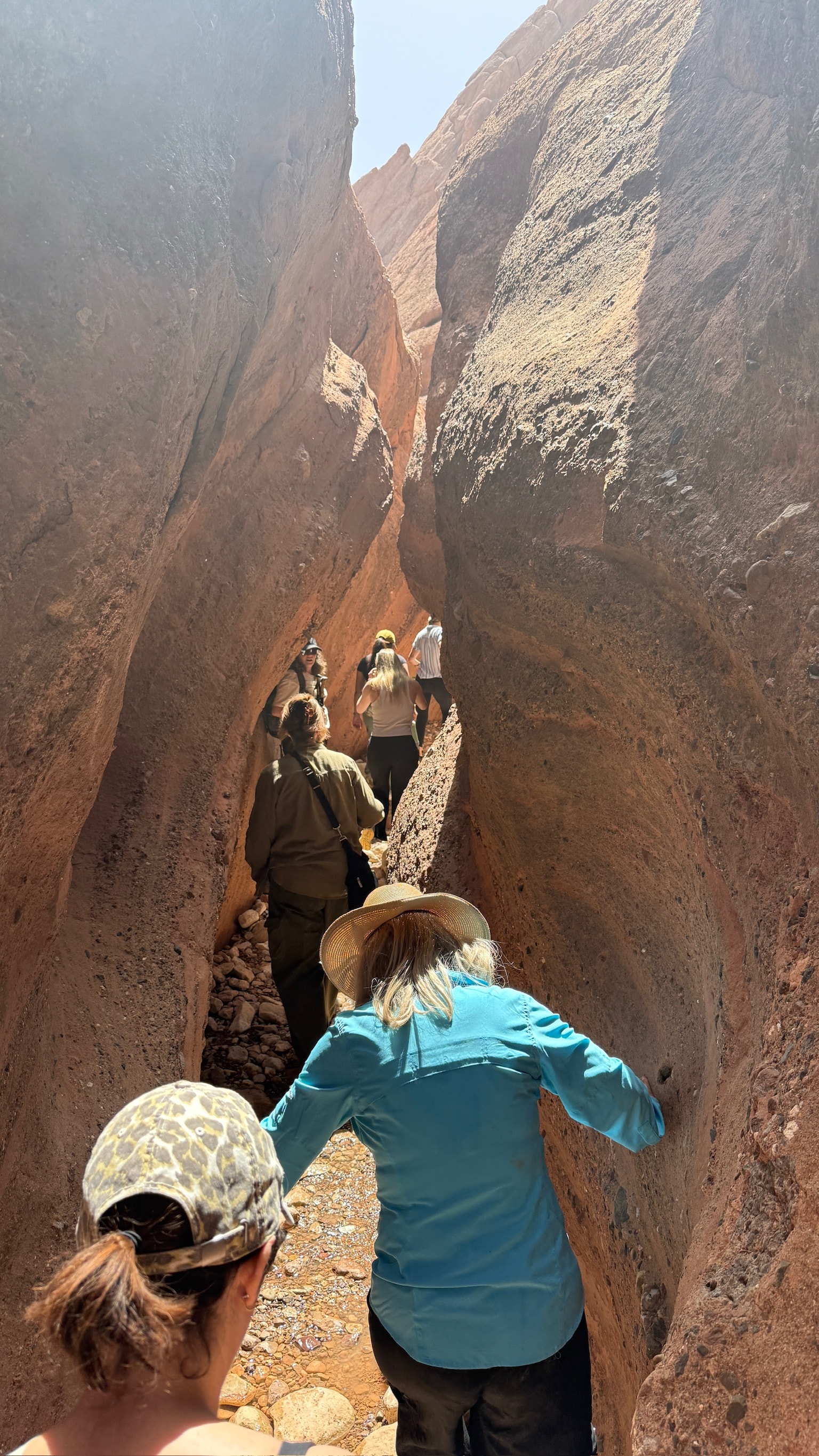The group squeezing single-file through a narrow red rock slot canyon in the Atlas Mountains, hands braced against the walls on either side