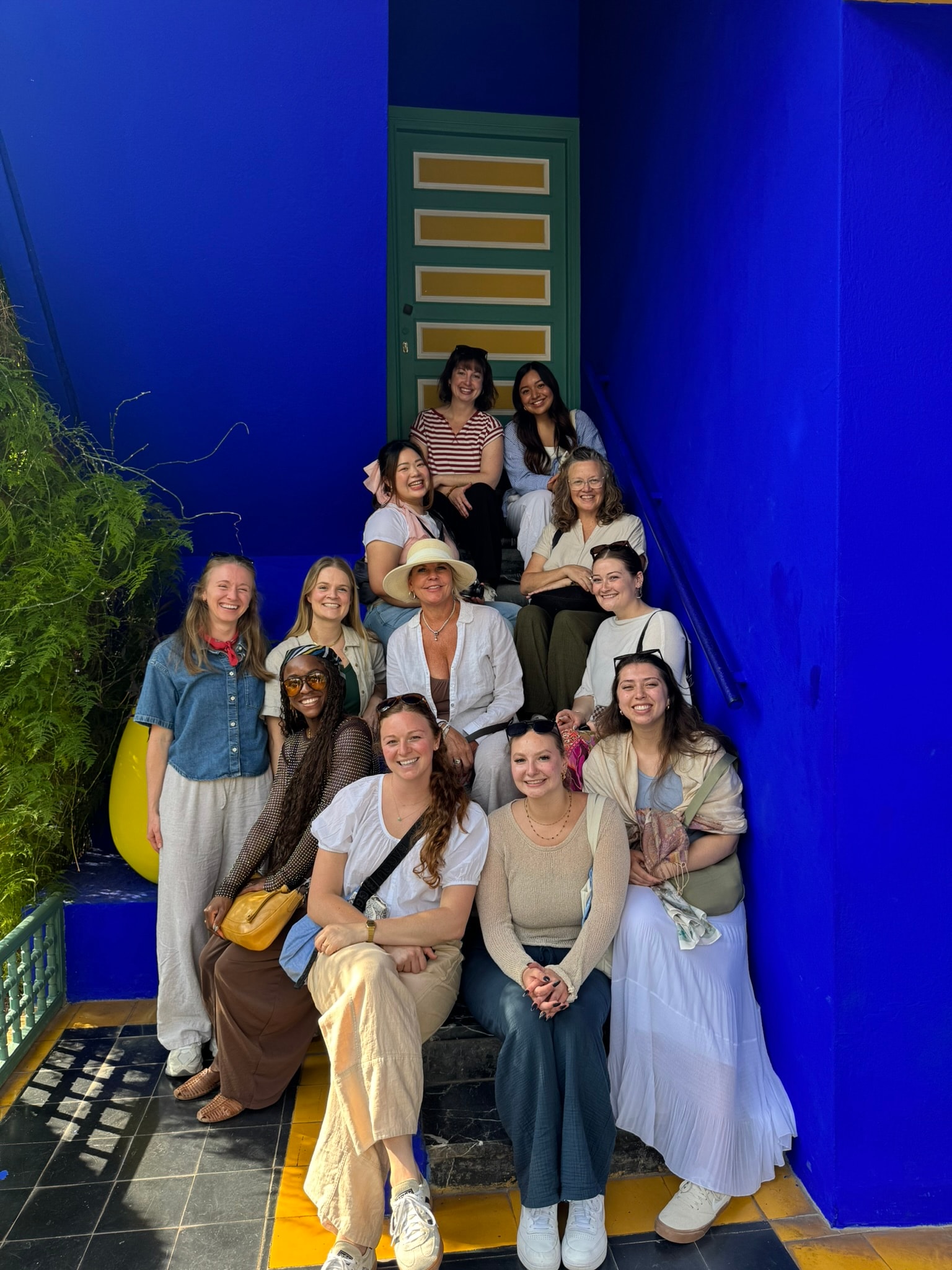 Eleven women from the Morocco group posing together on the cobalt blue steps of Jardin Majorelle in Marrakech, laughing and smiling