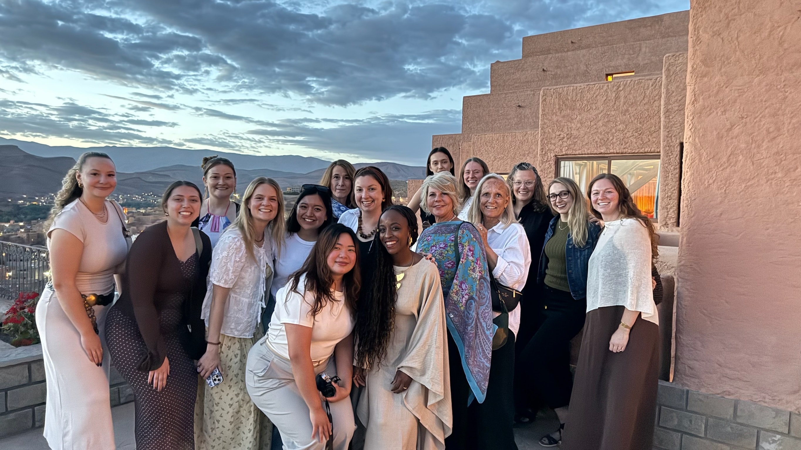 All 16 women on a rooftop terrace at dusk, Atlas Mountains and a dramatic cloudy sky stretching behind them, Morocco